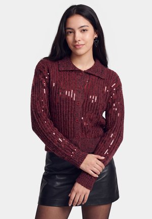 Ung kvinde med langt mørkt hår iført en burgundy strikket cardigan med pailletter og en sort lædernederdel, stående foran en ensfarvet baggrund.