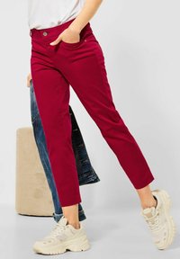 Red cotton blend pants with a slim fit, side pockets, and a button closure; paired with a light top and beige sneakers.