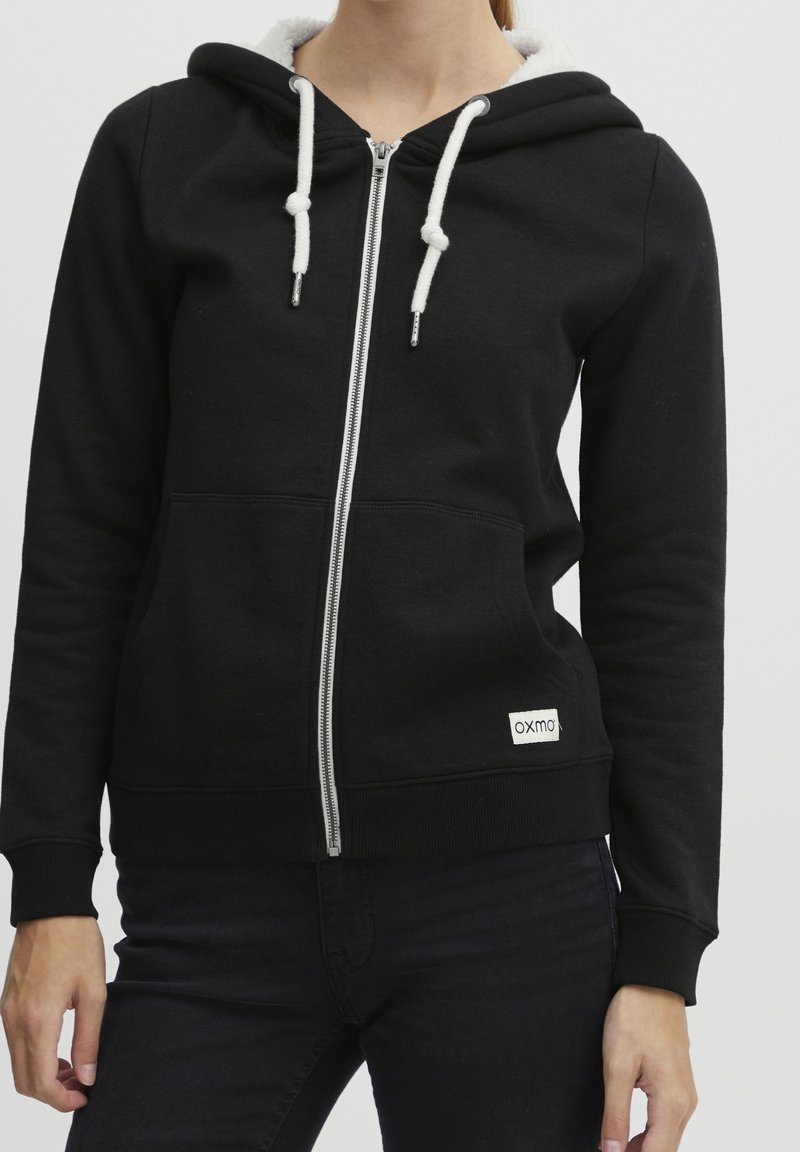 Personne portant un sweat à capuche noir zippé avec des cordons blancs et une capuche doublée en polaire, associée à un pantalon sombre.