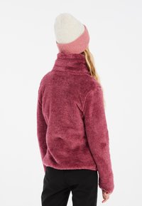 Fuzzy vinröd fleecjacka med hög krage och långa ärmar, tillsammans med en ljusfärgad mössa med en rosa band. Svarta byxor synliga.