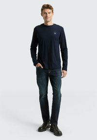 Granatowa bawełniana koszulka z długim rękawem, z małą naszywką z logo na piersi, w zestawieniu z ciemnymi dżinsami i czarnymi botkami.