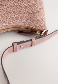 Sac à main rose tissé texturé avec fermeture éclair dorée et bandoulière assortie réglable sur une surface blanche.