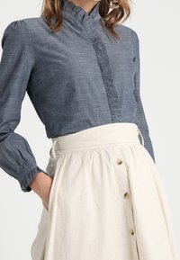 Chemise en chambray bleu avec un col volants et des boutons, associée à une jupe en lin crème à taille haute avec des boutons dorés.