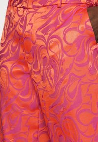 Pantalon taille haute orange et rose avec des motifs abstraits tourbillonnants et une ceinture à passants, porté avec une main dans la poche.