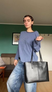 Bolso tote de cuero negro con detalle de costura vertical y dos asas largas, sostenido por una persona que lleva una camisa de rayas azul y blanca de manga larga y jeans holgados.
