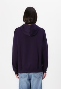 Felpa viola con cappuccio e maniche lunghe, caratterizzata da una vestibilità rilassata e polsini a costine. Cuciture visibili sul retro; indossata con jeans azzurri chiari.