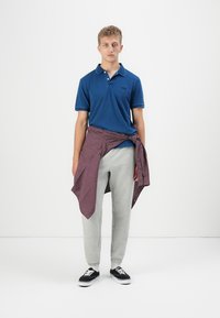 Modrá polo tričko, sivé tepláky a červená károvaná košeľa viazaná okolo pása. Čierne tenisky a biele ponožky dotvárajú outfit.