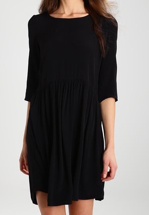 Woman wearing a loose-fitting black dress with three-quarter sleeves and a gathered waist, standing against a plain light background.