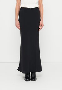 Jupe noire longue jusqu'au sol, à texture lisse, taille haute, avec un léger évasement à l'ourlet. Portée avec des bottines noires.