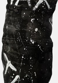 Tissu en denim noir avec un motif de éclaboussures blanches, présentant des accents métalliques subtils. Gros plan mettant en valeur la texture et les détails de couture.