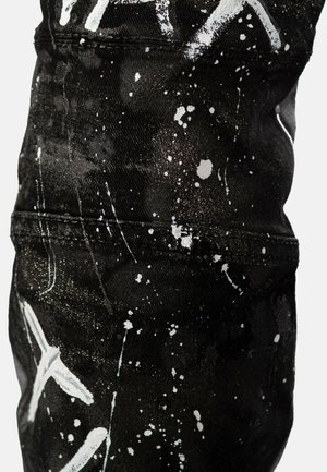 Zwarte denimstof met een wit spetterpatroon, voorzien van subtiele metallic accenten. Close-upweergave die de textuur en stikdetails benadrukt.