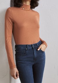 Långärmad polotröja i rostorange, med en mjuk textur och guldfärgade detaljer. Kombinerad med mörkblå högmidjade jeans.