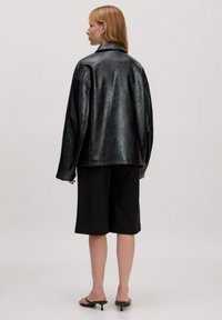Zwarte neplederen oversized jasje met een kraag, verlaagde schouders en een gladde textuur, gecombineerd met losse zwarte shorts.