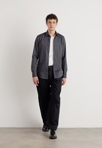 Graues Langarmhemd mit Knopfdesign, über einem weißen T-Shirt getragen, kombiniert mit schwarzen Hosen und schwarzen Schuhen. Schlichter Hintergrund.