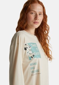 Eine rothaarige Frau trägt ein cremefarbenes Langarmshirt mit einer Grafik von Minnie Mouse und dem Text "Nimm es leicht, Minnie und Freunde."