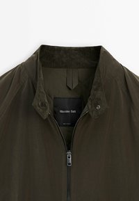 Olijfgroene bomberjack met een geribbelde kraag, ritssluiting en doorgestikte details. Label met "Massimo Dutti", gemaakt in Vietnam.