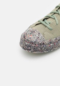 Sneaker vert avec une tige en maille texturée et un capuchon en caoutchouc gris marbré avec des éclaboussures colorées. Lacets verts assortis.
