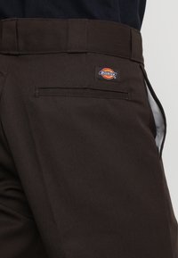 Pantalones de trabajo marrones con corte recto, fabricados en una tela duradera, que cuentan con un bolsillo trasero y un pequeño parche con el logo de Dickies en la cinturilla.