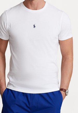 Mann trägt ein schlichtes weißes kurzärmeliges T-Shirt mit kleinem marineblauem Polospieler-Logo und blauer Hose, Hände in den Taschen, Armbanduhr sichtbar.