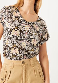 Černý top s barevnými květinovými vzory a krátkými rukávy, zkombinovaný se světle béžovými kalhotami s vysokým pasem a knoflíkovými detaily.