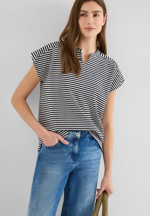 Jonge vrouw die een zwart-wit gestreept T-shirt met korte mouwen en een blauwe spijkerbroek draagt, met één hand in de zak en een olijfgroene tas vasthoudend.