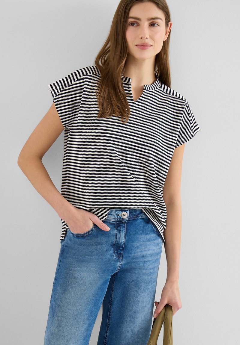 Jeune femme portant un haut à manches courtes rayé noir et blanc et un jean bleu, une main dans la poche et tenant un sac vert olive.