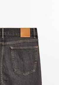 Jeans in denim nero con una toppa in pelle marrone chiaro sulla parte posteriore della vita e dettagli di cuciture visibili; presenta due tasche posteriori.