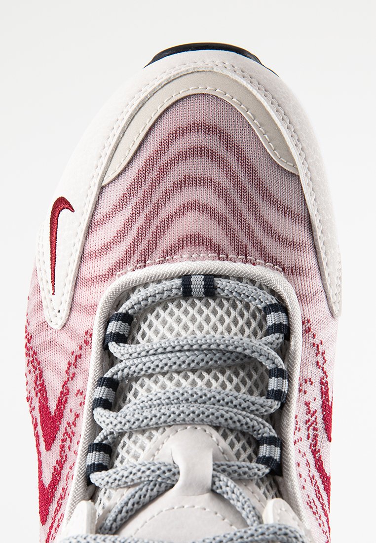 Sneaker con una toma in rete bianca che presenta un motivo a onde rosse, lacci grigi e una punta rinforzata. Dettagli del logo rosso vicino all'area dei lacci.