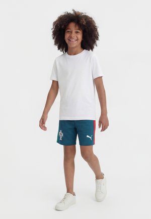 Naeratav laps lokkis juustega, valges t-särgis, sinistes Portugali jalgpallipükstes ja valgetes tennistes, kõndimas tavalise tausta ees.