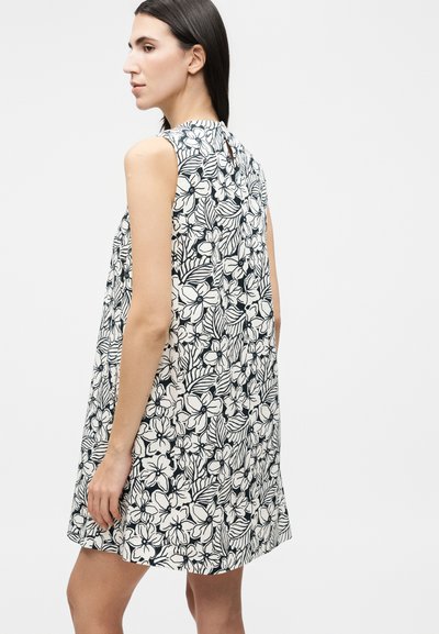 Γυναίκα με αμάνικο ασπρόμαυρο φόρεμα με ανθικό μοτίβο, στραμμένη προς τα αριστερά, σε απλό λευκό φόντο.