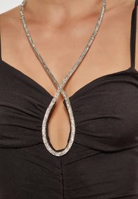 Colier din strasuri cu formă de picătură, de culoare argintie, cu un design împletit, purtat peste un top dintr-un material negru ajustat, cu detalii de strângere.