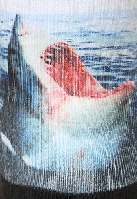 Stickade strumpor med en fiskdesign i nyanser av blått, rött och vitt mot en texturerad, flerfärgad bakgrund med horisontella ränder.