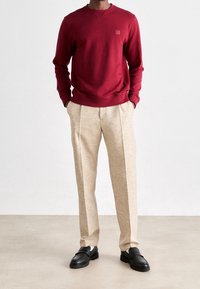 Sweat-shirt rouge à col rond et poignets côtelés, associé à un pantalon beige tailleur à finition texturée et des chaussures noires.