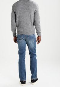 Pull en tricot gris, jean bleu en denim, vue de dos ; coupe décontractée avec tissu texturé, poignets côtelés et design simple. Baskets noires aux pieds.