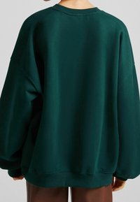 Sudadera verde de gran tamaño en tejido suave con cuello redondo y hombros caídos. Puños y dobladillo de canalé; textura lisa, diseño minimalista.