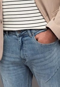 Los jeans de mezclilla azul claro presentan un diseño clásico de cinco bolsillos, un cierre con botón de metal y un ligero desgaste para añadir textura. Se llevan con una camiseta a rayas.