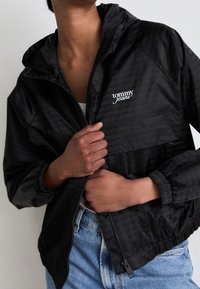 Svart vindjacka med huva, med en subtil glansig yta och "tommy jeans"-logotyp på bröstet. Dragkedjestängning, elastiska mudd.
