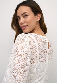 Hvid blonde-top med lange ærmer, blomstermønstre, en nøglehul-design på ryggen og dekorative knapper langs ryggen. Glat tekstur.
