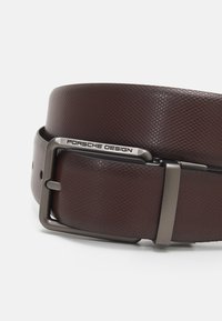 Cinturón de cuero texturizado marrón con hebilla de metal gris que presenta un grabado "Porsche Design". El diseño destaca la durabilidad y el estilo.