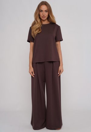 Kobieta o długich blond włosach, ubrana w luźną ciemnobrązową koszulkę i szerokie spodnie, stojąca na jednolitym tle.