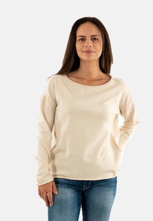 Donna con lunghi capelli castani, che indossa una maglietta crema a maniche lunghe e jeans blu, in piedi con una mano in tasca su uno sfondo semplice.