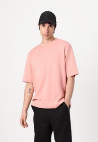 Ροζ κοντομάνικο μπλουζάκι από μαλακό ύφασμα; στρογγυλή λαιμόκοψη; χαλαρή εφαρμογή; σε συνδυασμό με μαύρα παντελόνια; μινιμαλιστικός σχεδιασμός, χωρίς αξιοσημείωτα μοτίβα.