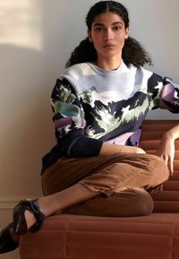 Strikket sweater med et bjerglandskabsmønster i nuancer af blå, grøn og lila; ribbede navy manchetter; parret med brune fløjlbukser.