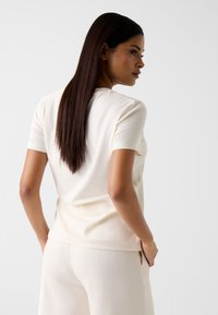 Kremowa bluzka z krótkim rękawem wykonana z miękkiego materiału, z okrągłym dekoltem i luźnym krojem, połączona z jasnymi, luźnymi spodniami.