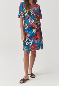 Kukkakuvioinen kietaisumekko, jossa sininen tausta ja moniväriset kukkakuviot. Lyhyet hihat ja polven mittainen malli. Mustat tasapohjaiset sandaalit.