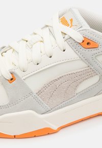 Detailní záběr na bílé a světle šedé tenisky Puma s oranžovými akcenty, texturovanými semišovými detaily a bílými tkaničkami.