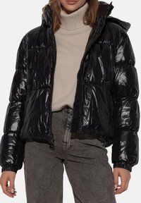 Giacca a vento nera con finitura lucida, dotata di cappuccio, chiusura con zip e polsini elastici. Indossata sopra un dolcevita beige e jeans grigi.