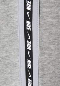 Šedá tkanina s texturovaným povrchem má černý sportovní pruh se opakovaným bílým značením "NIKE". Doplňky zahrnují kontrastní světle šedou.