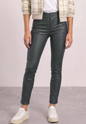 Femme portant un pantalon en cuir noir ajusté, des baskets blanches, une chemise blanche et une veste beige à carreaux, debout sur un sol gris.