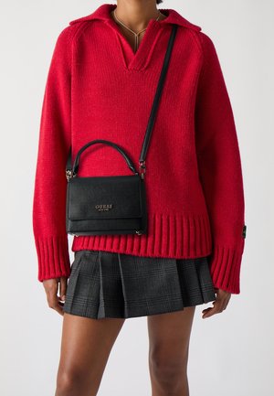 Personne portant un pull en maille rouge, une jupe plissée à carreaux gris et un sac bandoulière noir Guess avec logo doré.